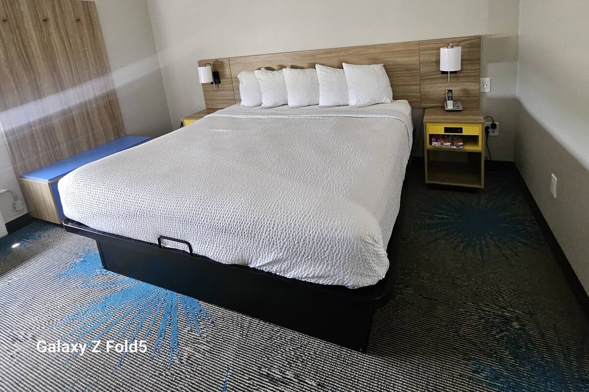 Ein Hotelzimmer mit Galaxy Z Fold5 Bett, einem Holz-Kopfteil, weißer Bettwäsche, einem Nachttisch mit Lampe und einer kleinen Ablage mit Gegenständen.
