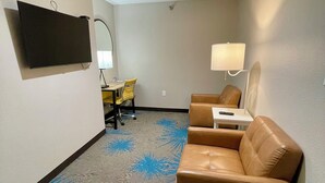 Habitación de hotel con sofá de cuero marrón, una mesita, una lámpara, un televisor montado en la pared y una alfombra con diseño azul.