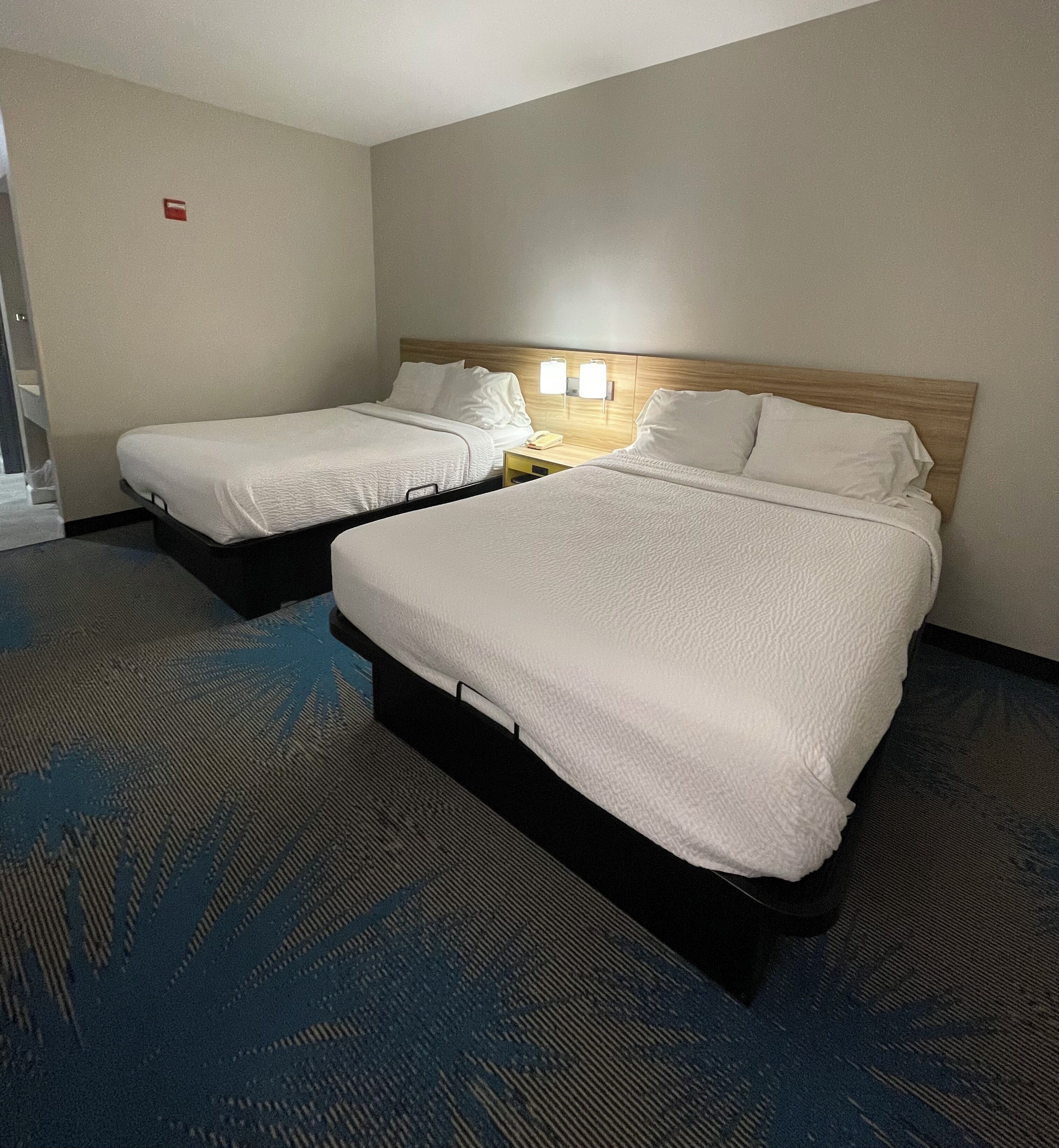 Habitación de hotel con dos camas, cabecera de madera, mesita de noche con lámpara y suelo alfombrado.