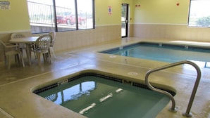 Una piscina interior con un pequeño jacuzzi, una mesa con sillas y una ventana que ofrece vistas a coches aparcados.