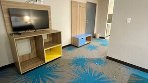 Habitación de hotel con televisión de pantalla plana montada en un estante de madera, un armario y una alfombra azul con diseño de hojas.