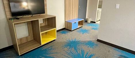 Ett hotellrum med en platt-TV som är monterad på en hylla i trä, en garderob och en blå matta med ett lövmönster.