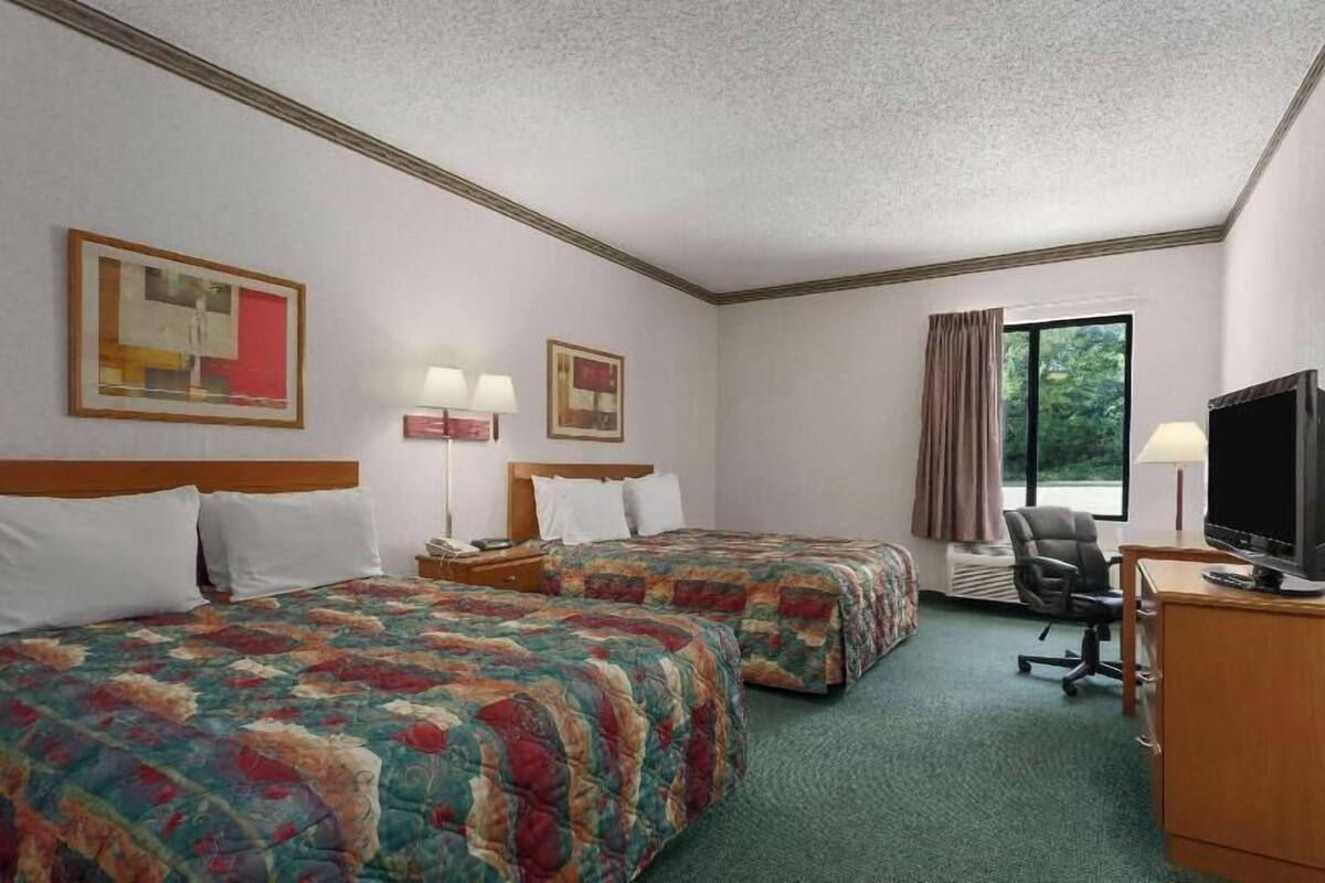 Ein Hotelzimmer mit zwei Betten, einem Schreibtisch mit Fernseher, einem Sessel, einem Fenster mit Vorhängen und Bildern an den Wänden.