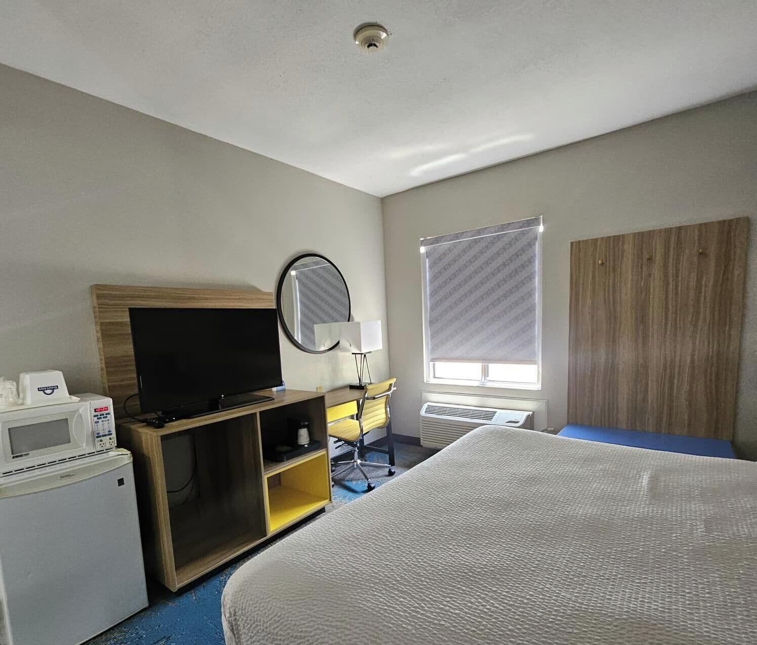 A hotel room with a Galaxy Z Fold5 bed, a flat-screen TV, a microwave, a small desk with a chair, a mirror, and a window with blinds.