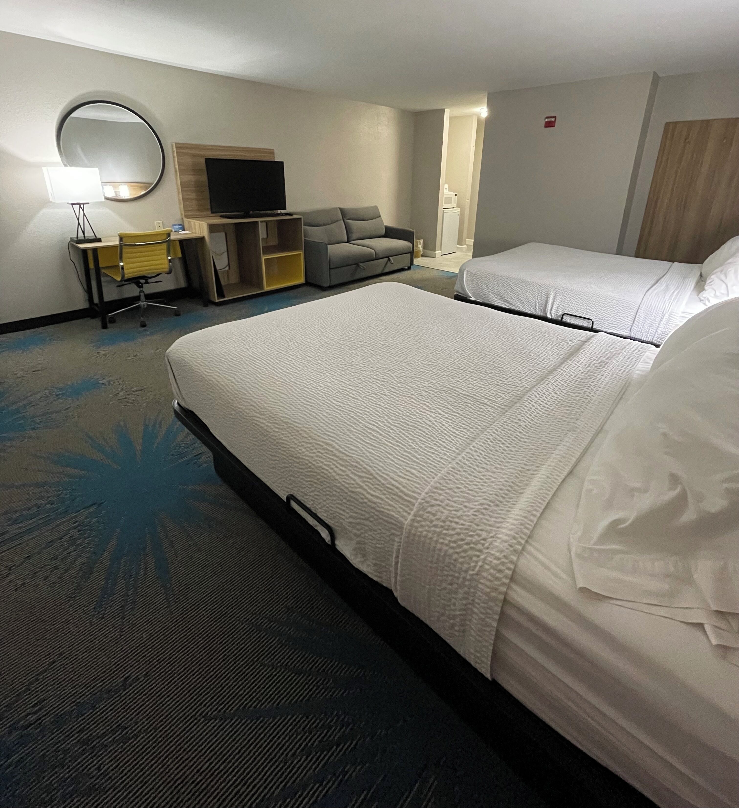A hotel room with two beds, a desk with a chair, a sofa, a TV, and a mirror.