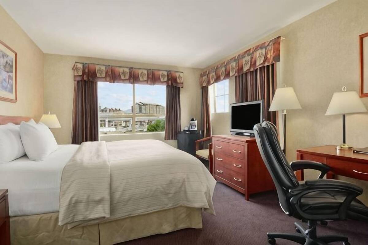 A hotel room with a bed, desk, chair, television, and a view of the city.