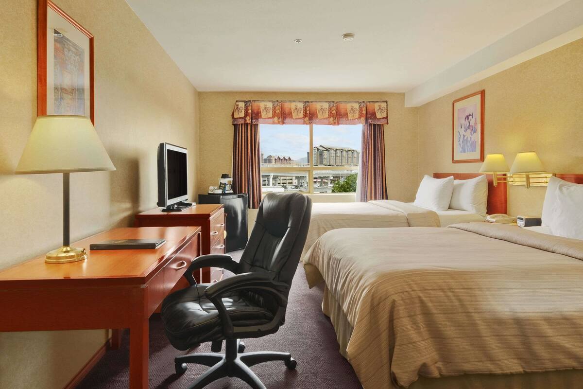 A hotel room with a desk, two beds, a chair, a TV, and a window with curtains.