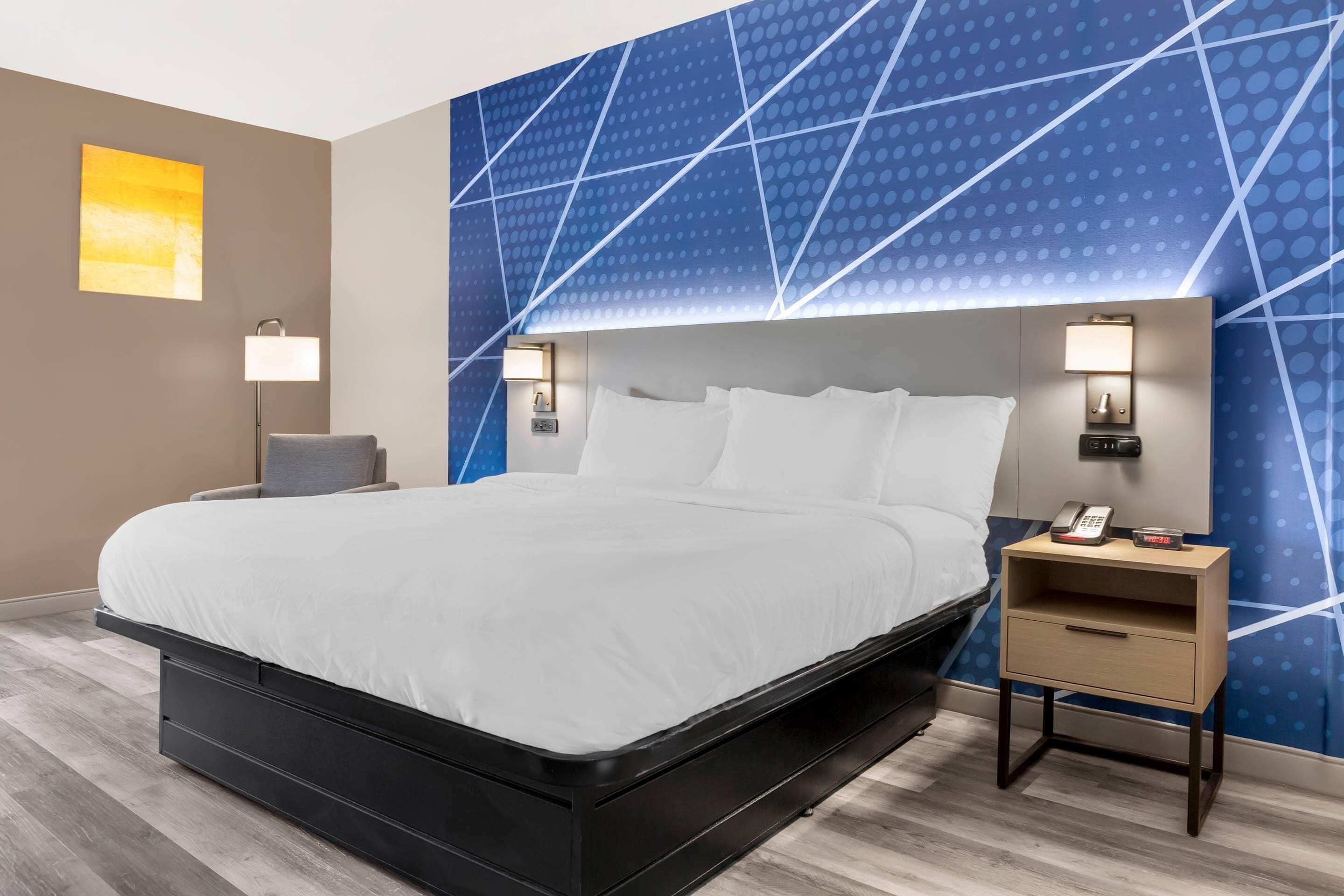 A modern hotel room with a large bed, bedside lamps, a nightstand, and a blue geometric wall design.