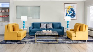 Un salon avec un canapé bleu, deux fauteuils jaunes, une table basse et un panneau indiquant « BIENVENUE ».