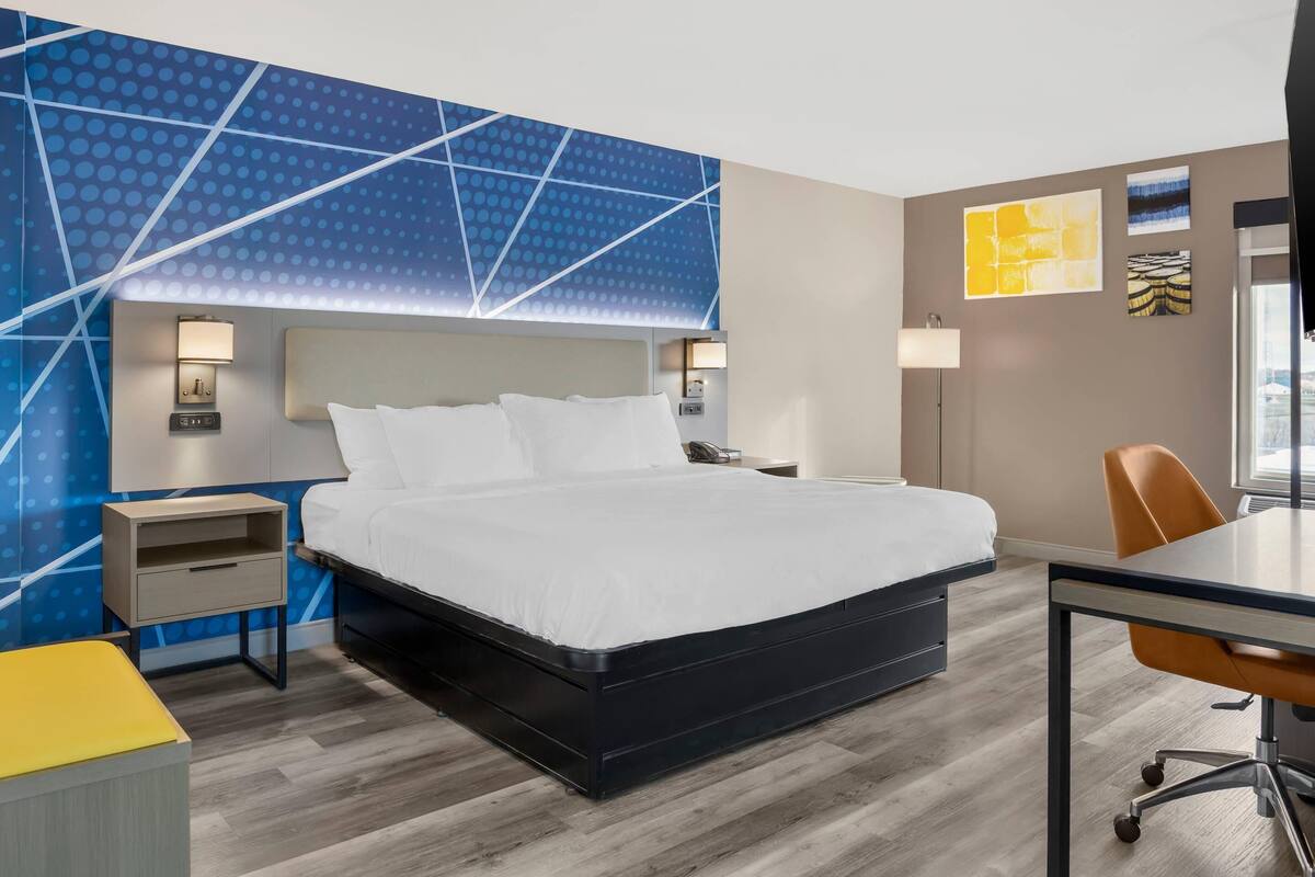 A modern hotel room with a large bed, bedside tables, a desk with a chair, and a view of the city.