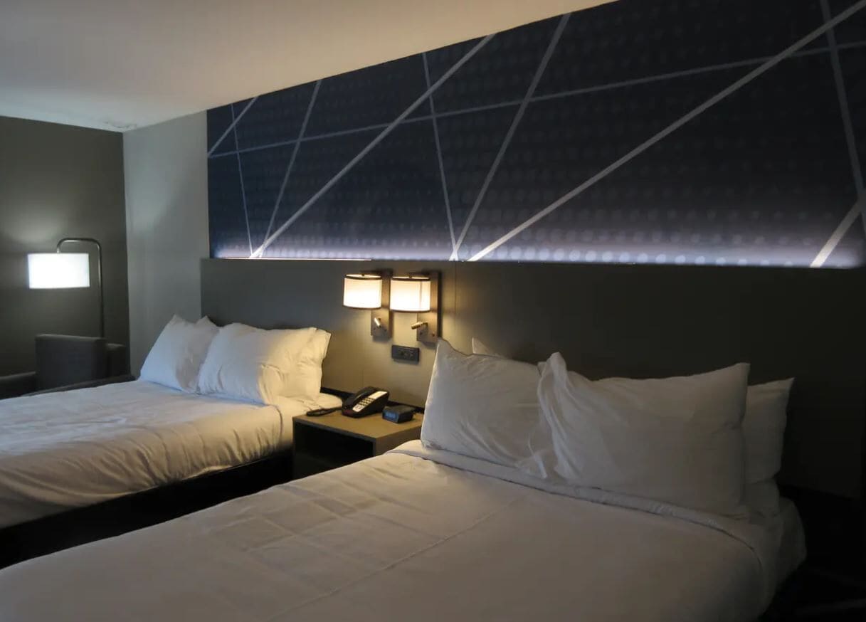 A hotel room with two beds, a nightstand with a lamp, and a wall-mounted light fixture.