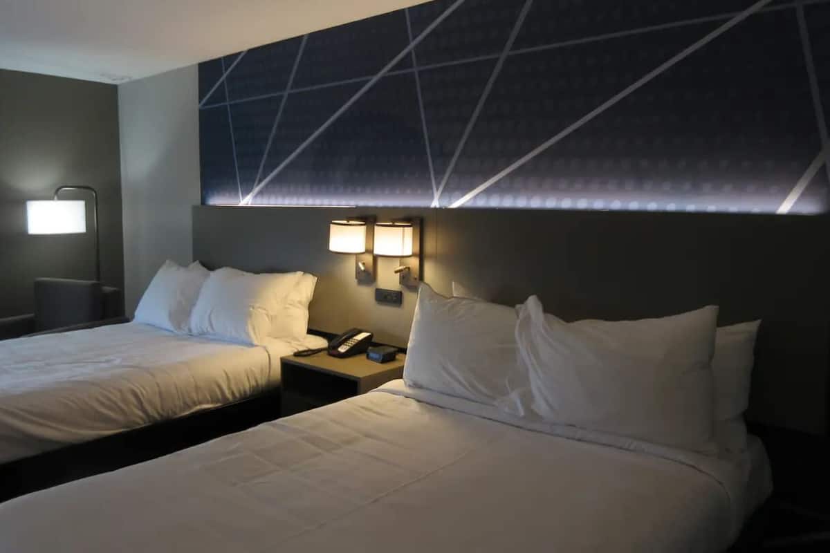 A hotel room with two beds, a nightstand with a lamp, and a wall-mounted light fixture.