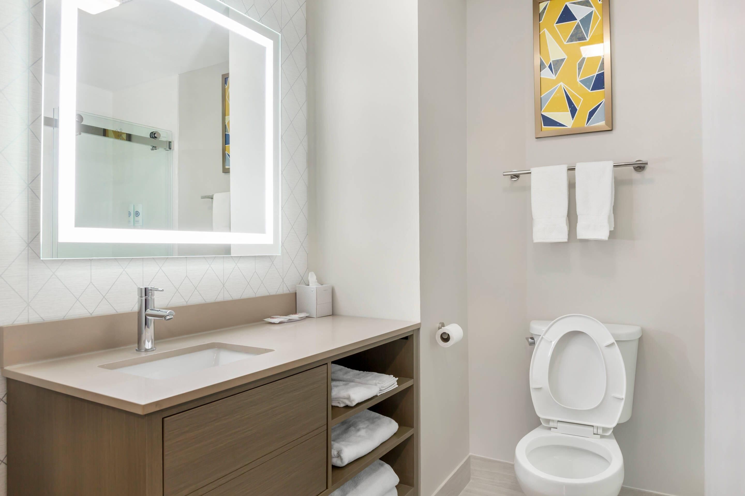 Une salle de bain équipée d’un lavabo, d’un miroir, d’un porte-serviettes, de toilettes et d’une œuvre d’art géométrique accrochée au mur.