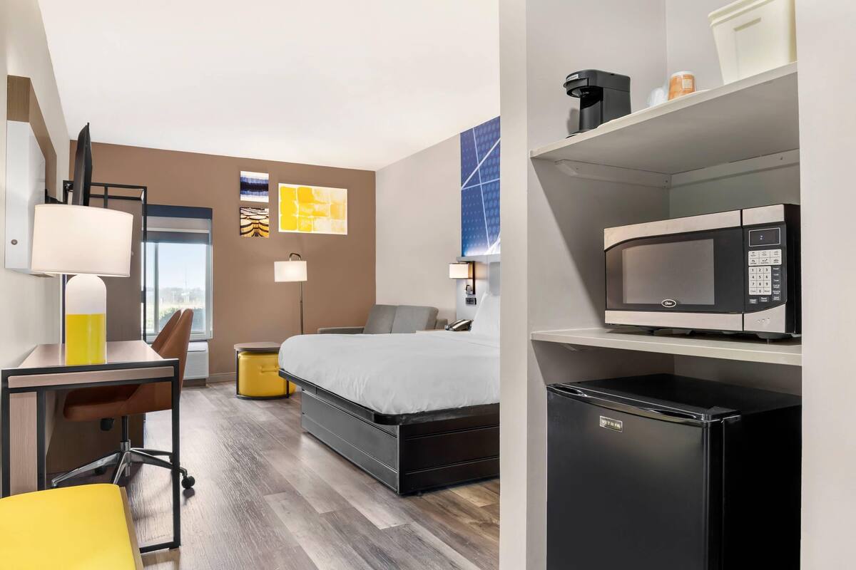 A hotel room with a bed, desk, chair, microwave, refrigerator, and a view of the city.