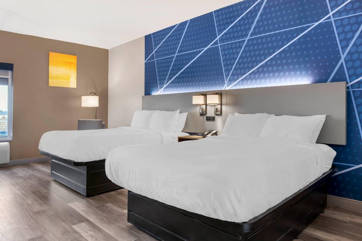 A hotel room with two beds, a large window, a wall with a geometric pattern, and a lamp.