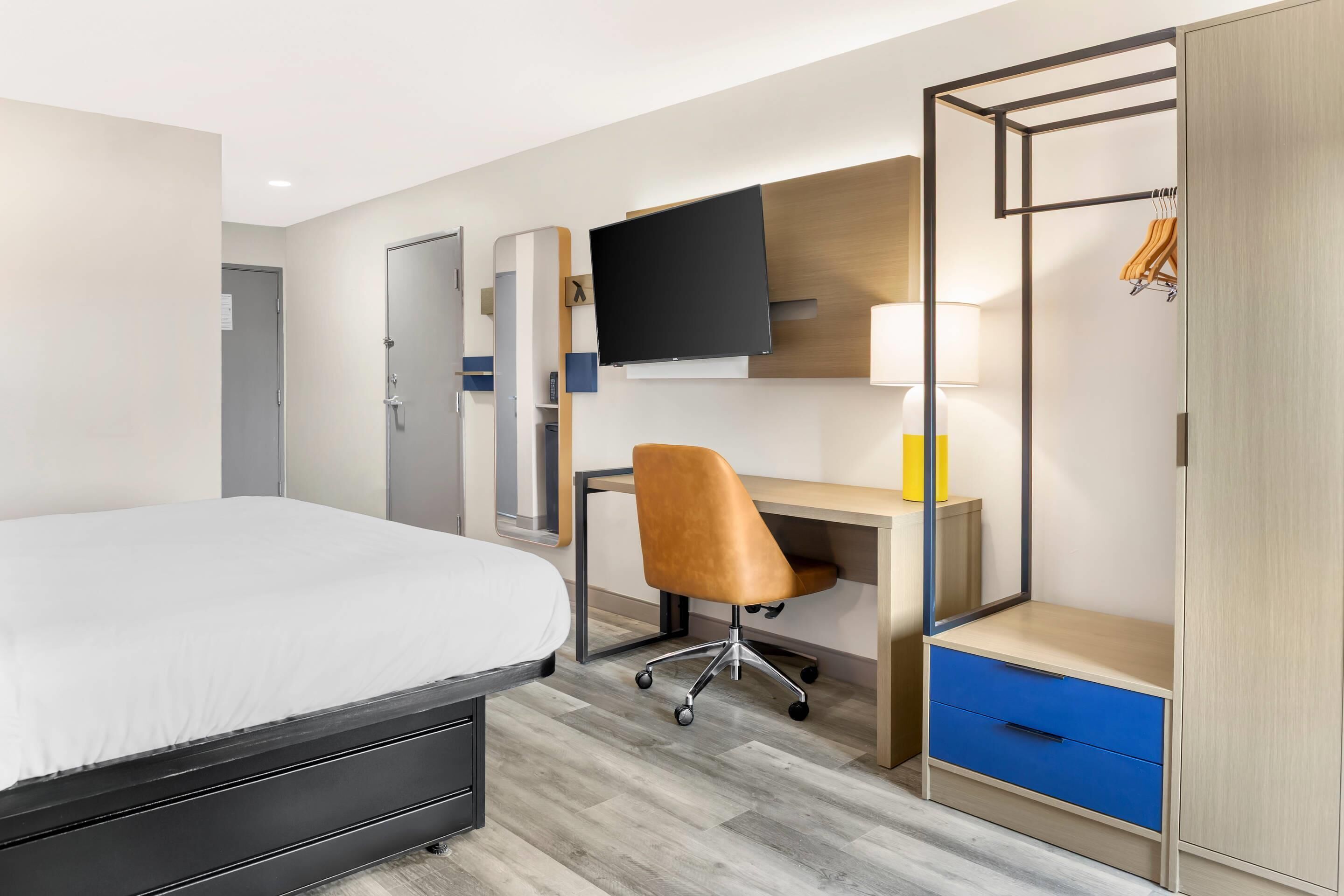 A hotel room with a bed, a desk, a chair, a TV, a wardrobe, and a lamp.