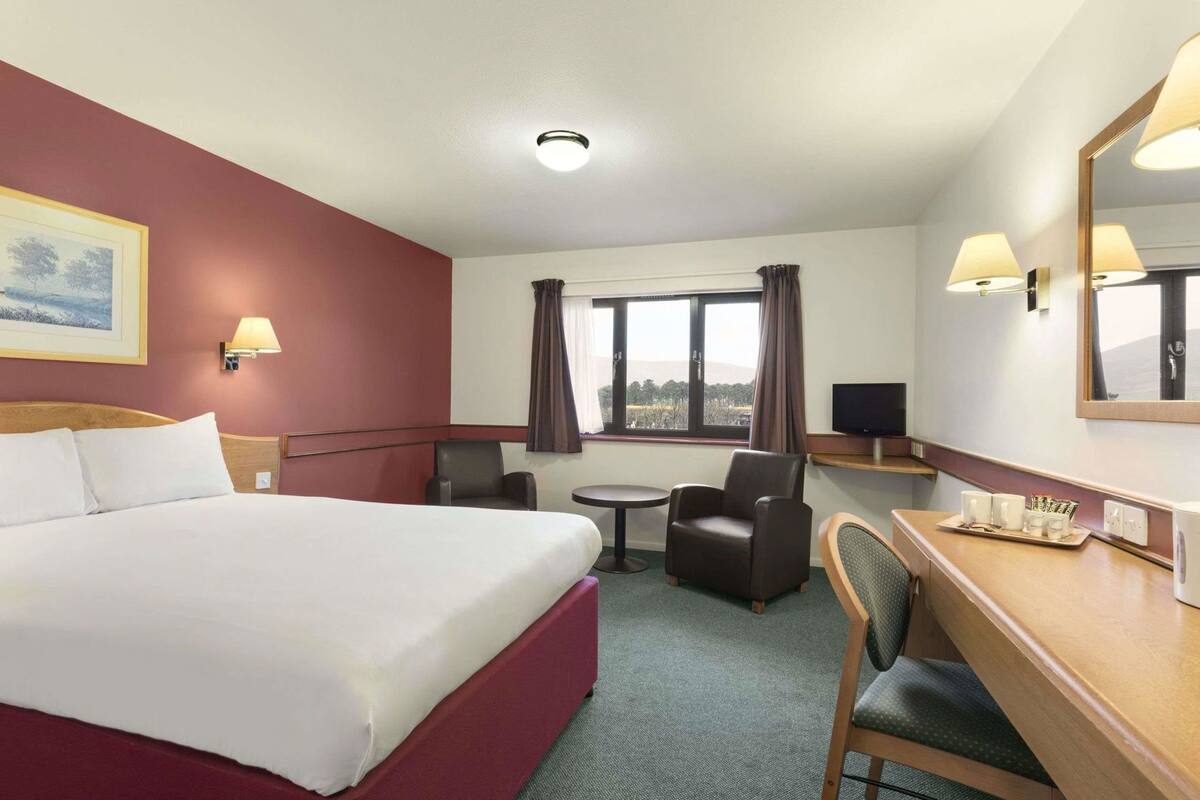 Ein Hotelzimmer mit einem Bett, einem Schreibtisch, einem Sessel, einem kleinen Tisch, einem Fernseher und einem Fenster mit Vorhängen.
