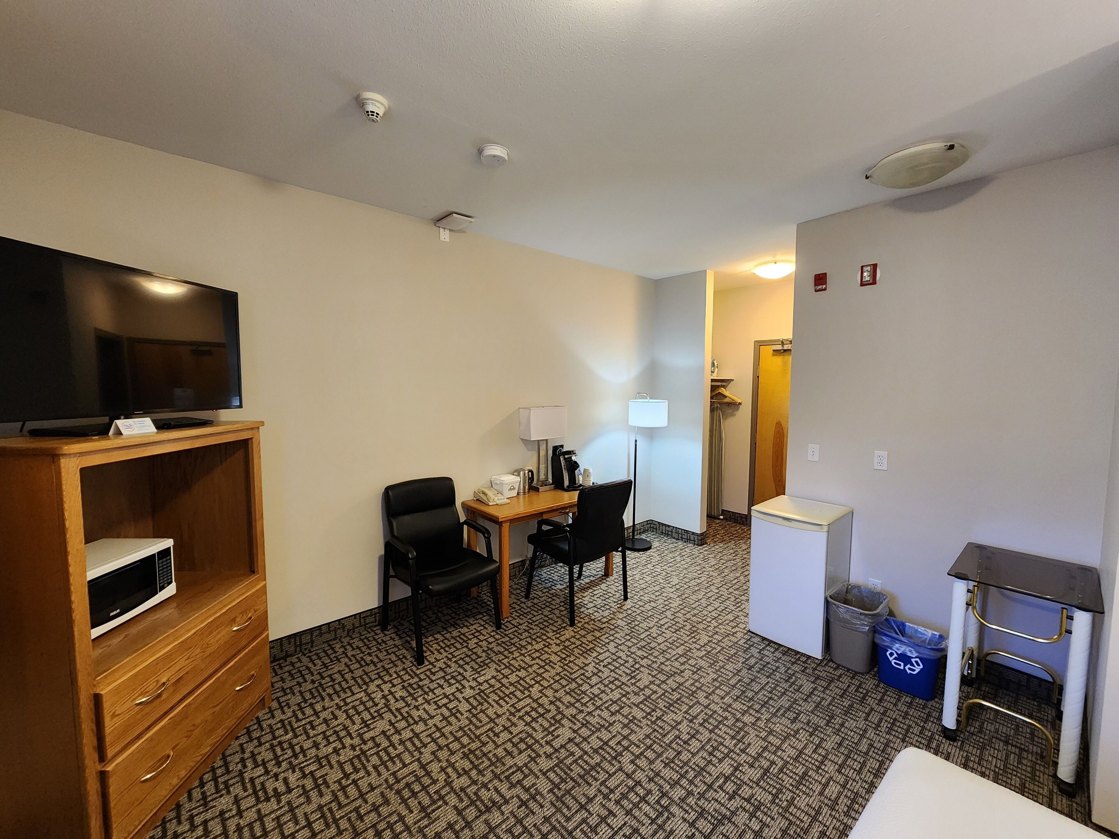 A hotel room with a flat-screen TV on a wooden stand, a desk with a lamp, two chairs, a small refrigerator, and a trash bin.