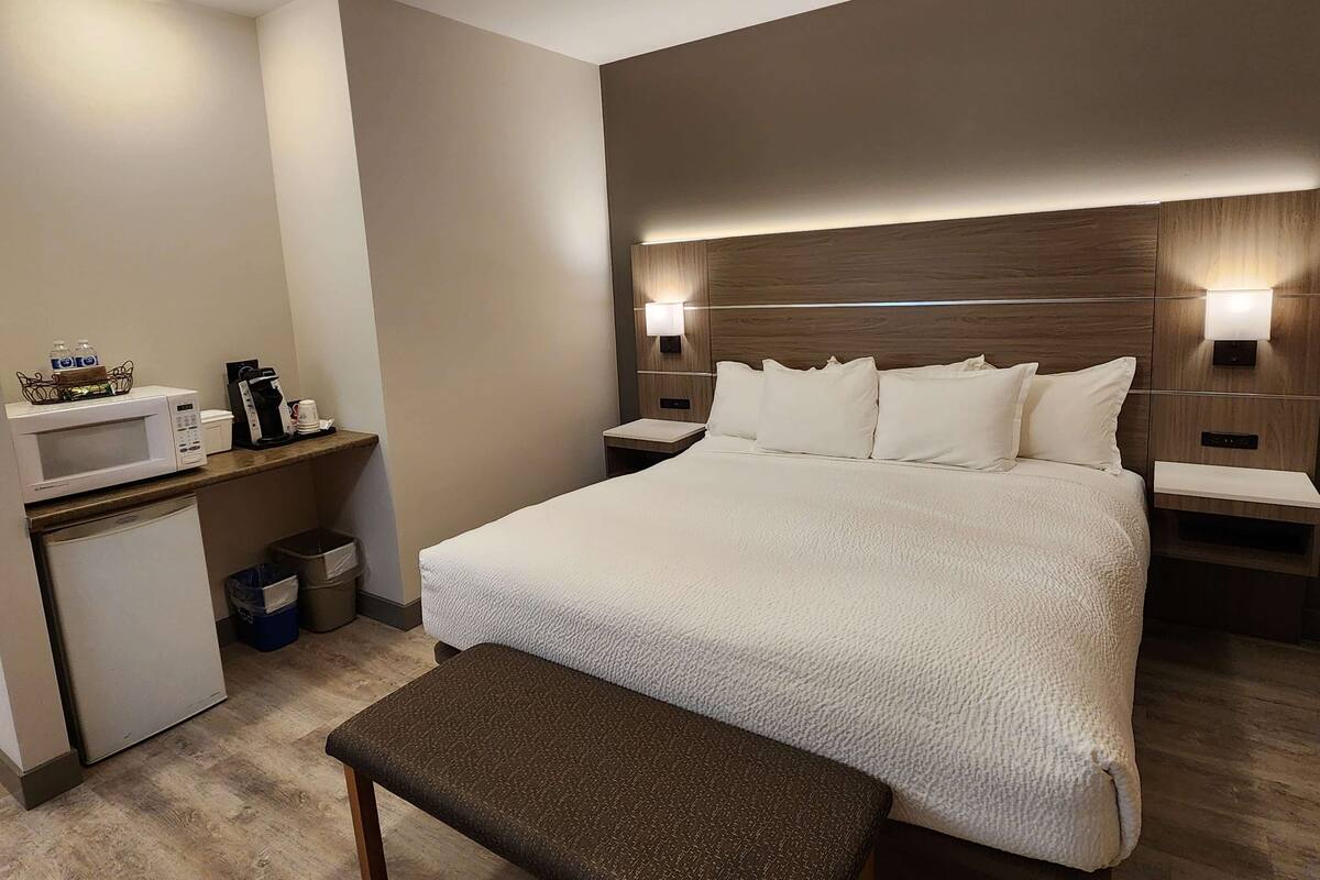 Habitación de hotel con una cama grande, mesitas de noche, microondas, refrigerador pequeño y un banco.