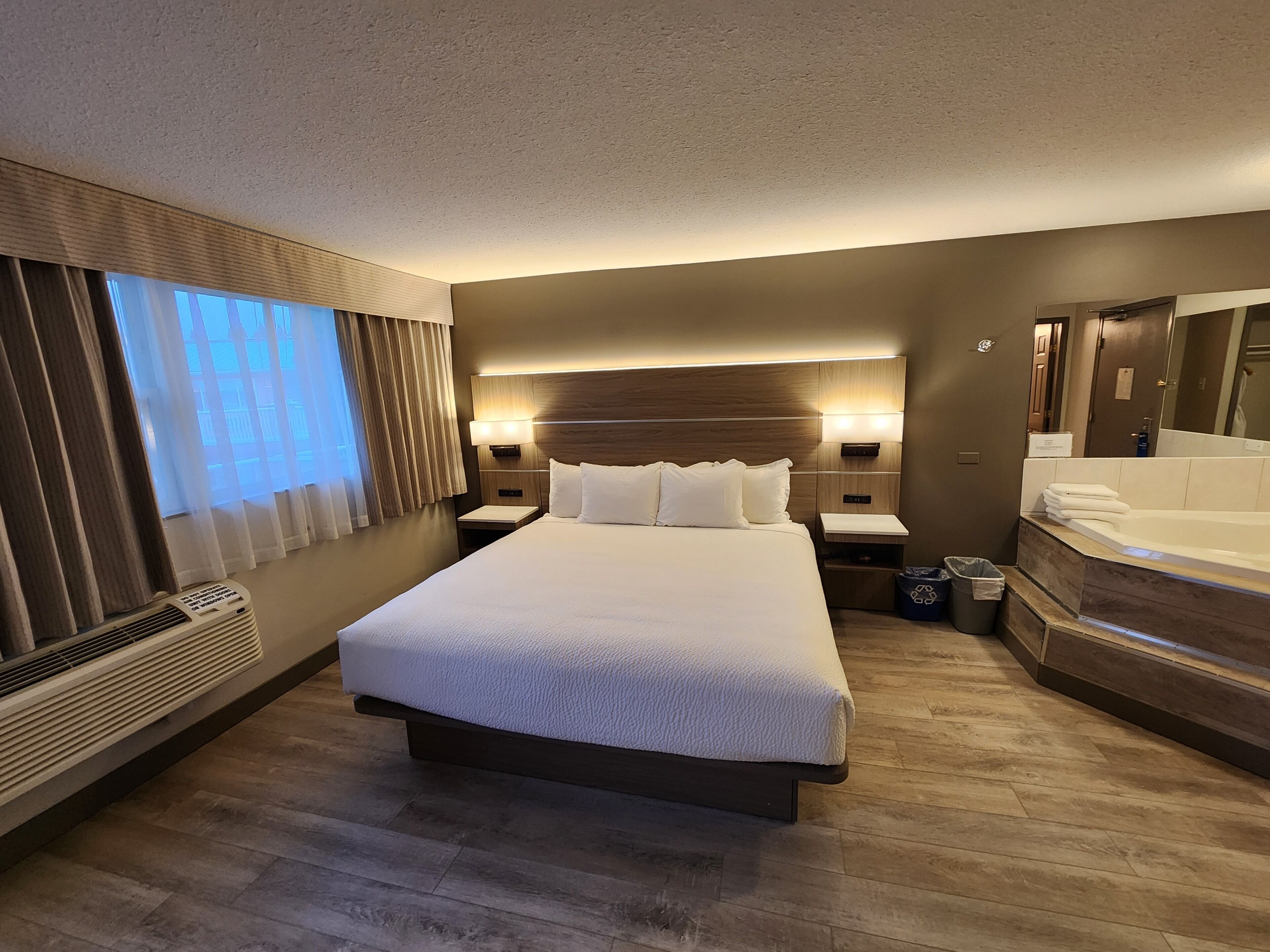 A hotel room with a large bed, bedside tables, a nightstand, a built-in bathtub, and a large window with curtains.