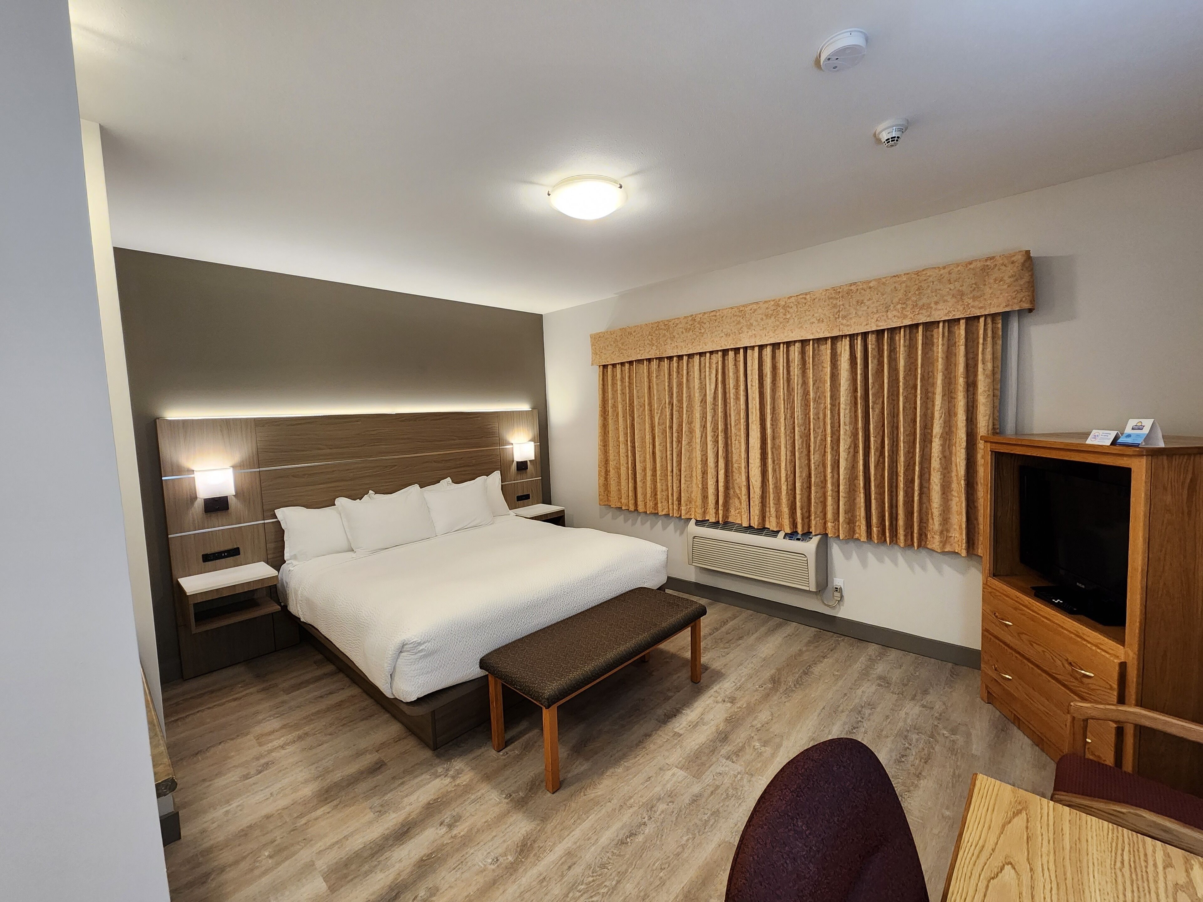 A hotel room with a bed, bedside tables, a bench, a TV cabinet, and a window with curtains.