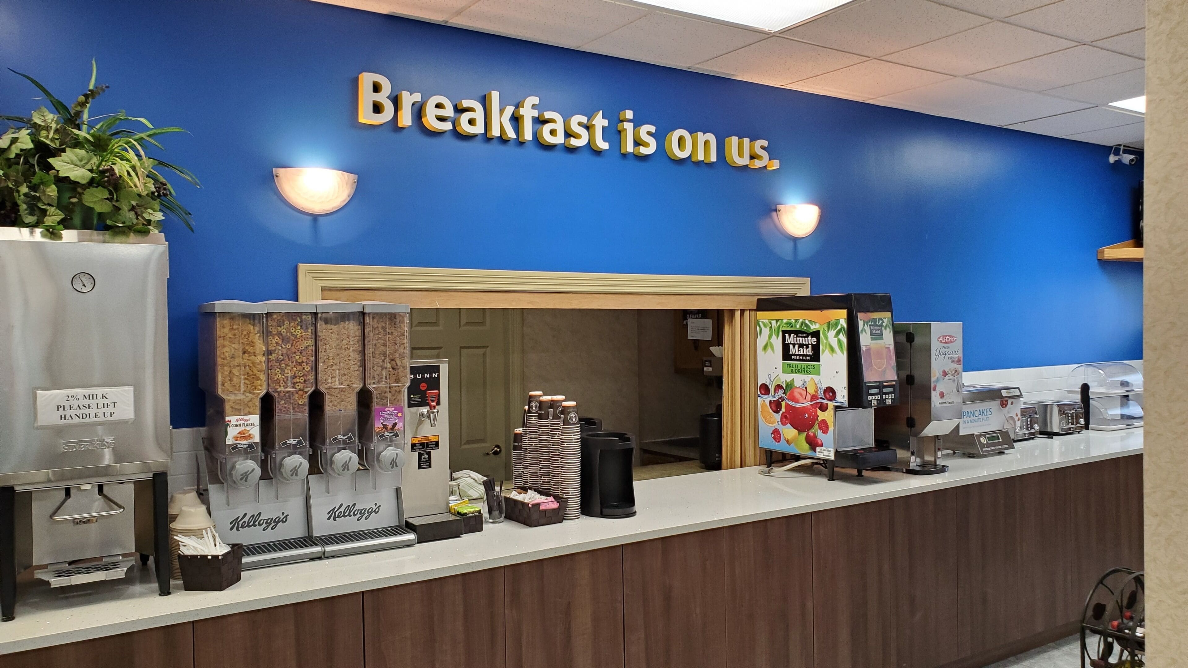 A breakfast counter with various coffee machines, a sign stating "Breakfast is on us," and a blue wall.