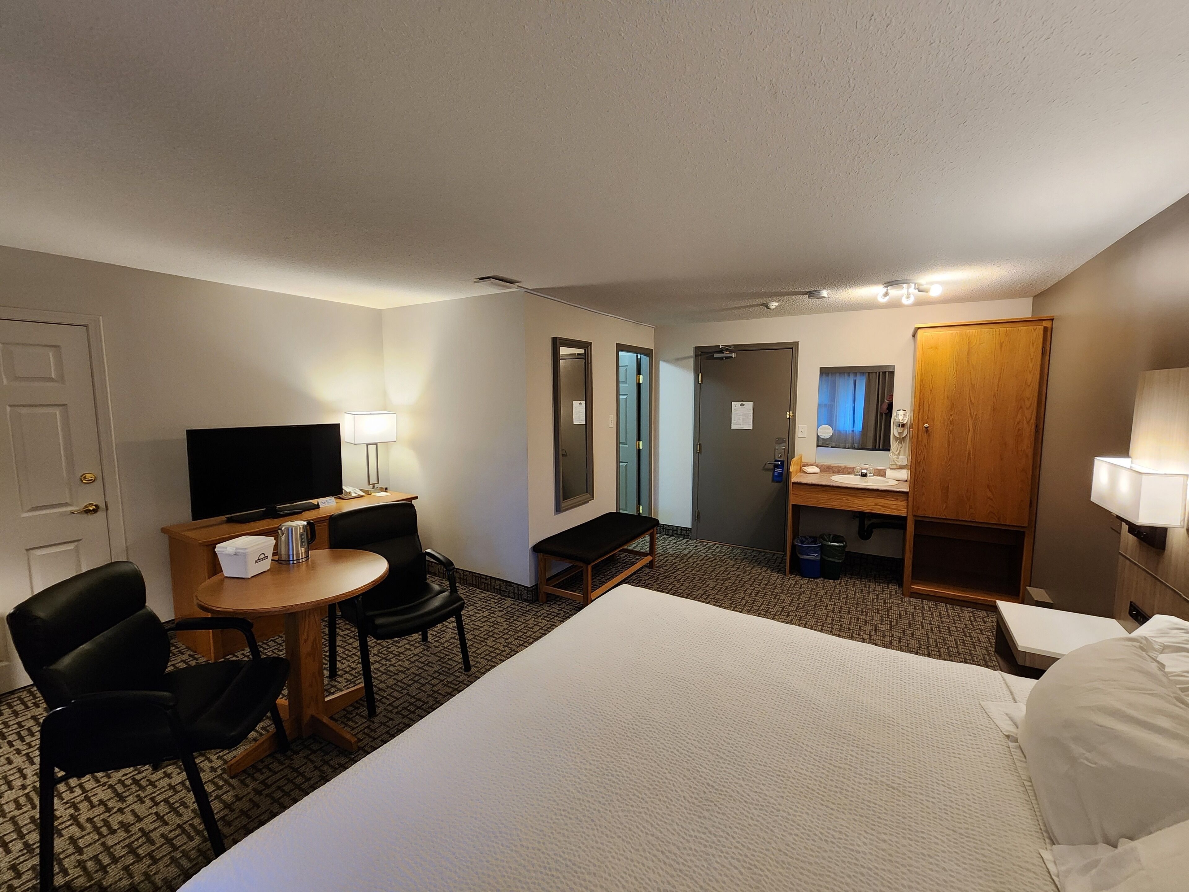 Habitación de hotel con cama, escritorio con silla, televisor, espejo, armario y baño con lavabo y espejo.