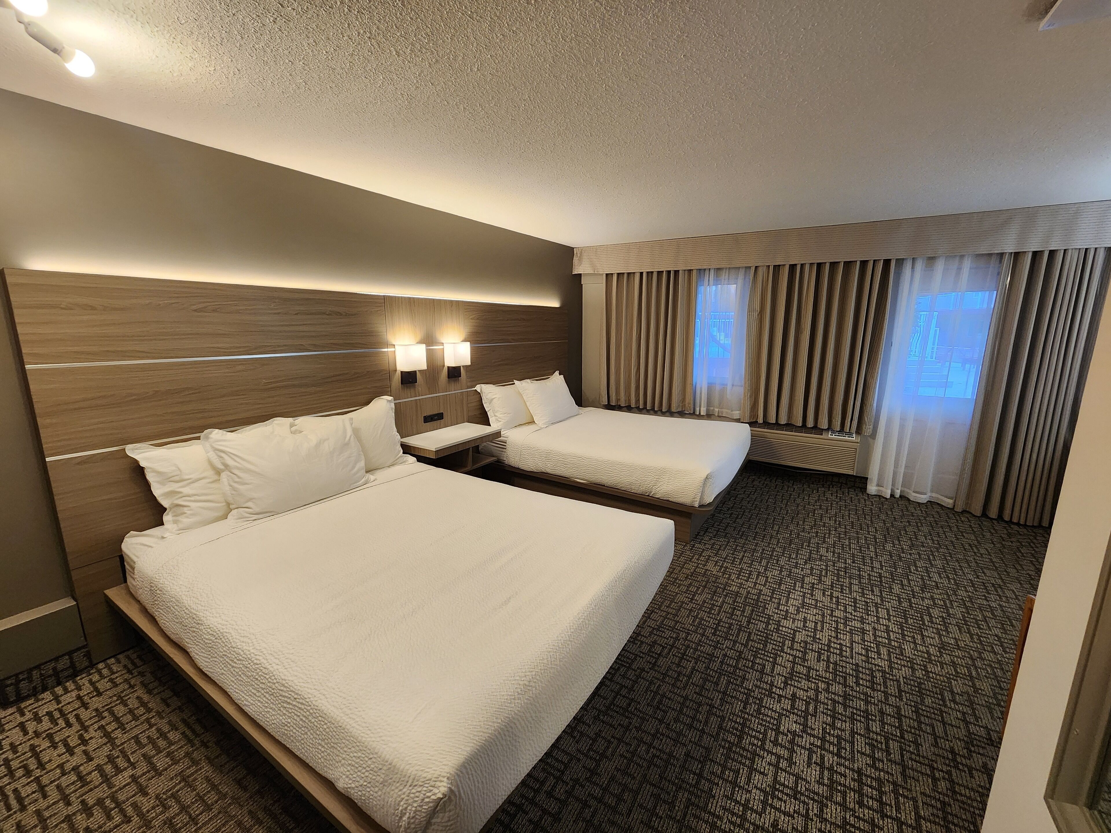 A hotel room with two beds, a nightstand, a small table, and a window with curtains.