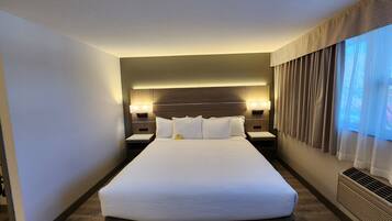 A modern hotel room with a large bed, two bedside tables with lamps, a built-in headboard, and a large window with curtains.