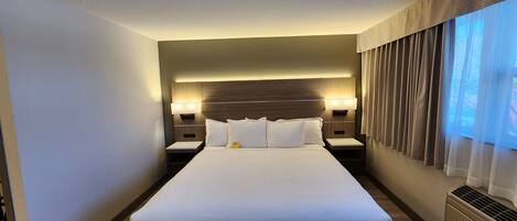 A modern hotel room with a large bed, two bedside tables with lamps, a built-in headboard, and a large window with curtains.