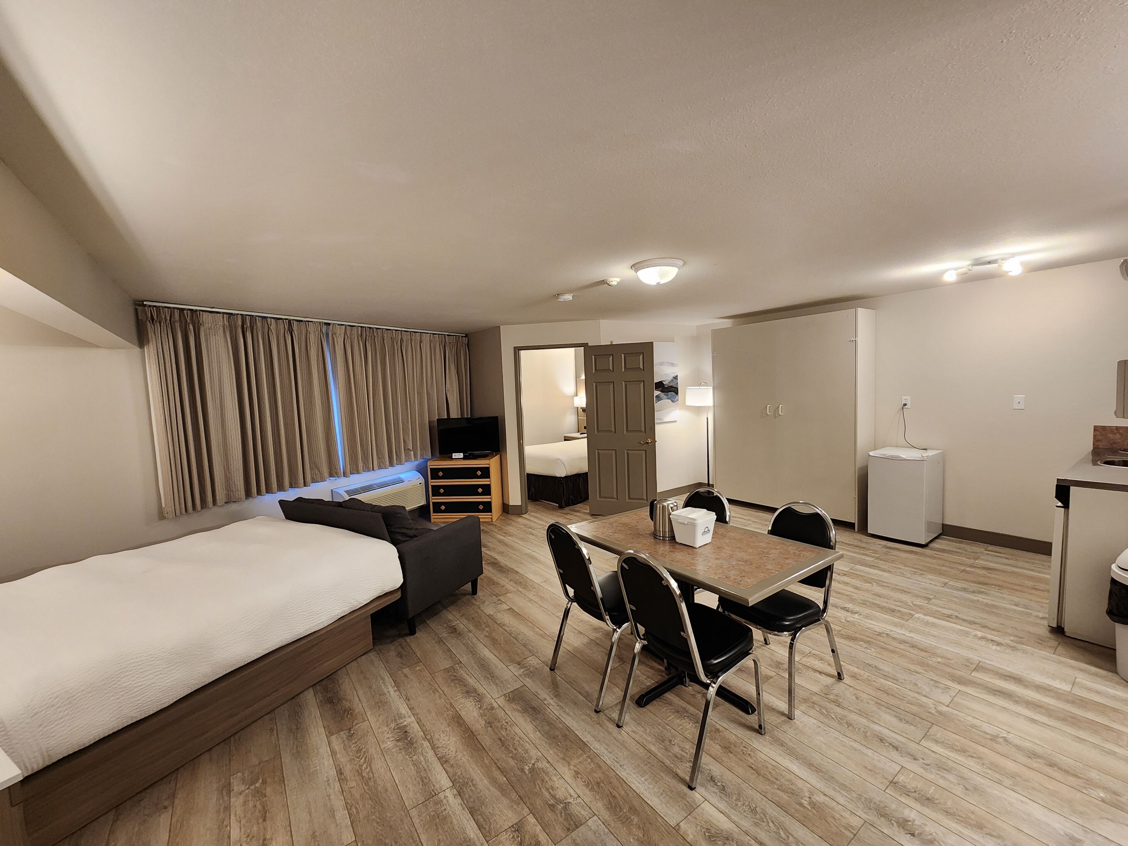 A modern hotel room with a bed, a dining table with chairs, a small kitchen area, and a separate bedroom.