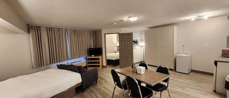 Quarto de hotel moderno com cama, mesa de jantar com cadeiras, pequena área de cozinha e quarto separado.