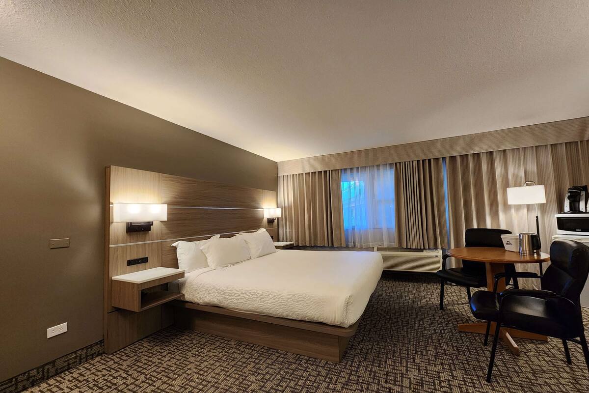 Habitación de hotel con una cama grande, mesitas de noche, un escritorio con una silla, una lámpara y vistas al exterior a través de cortinas.
