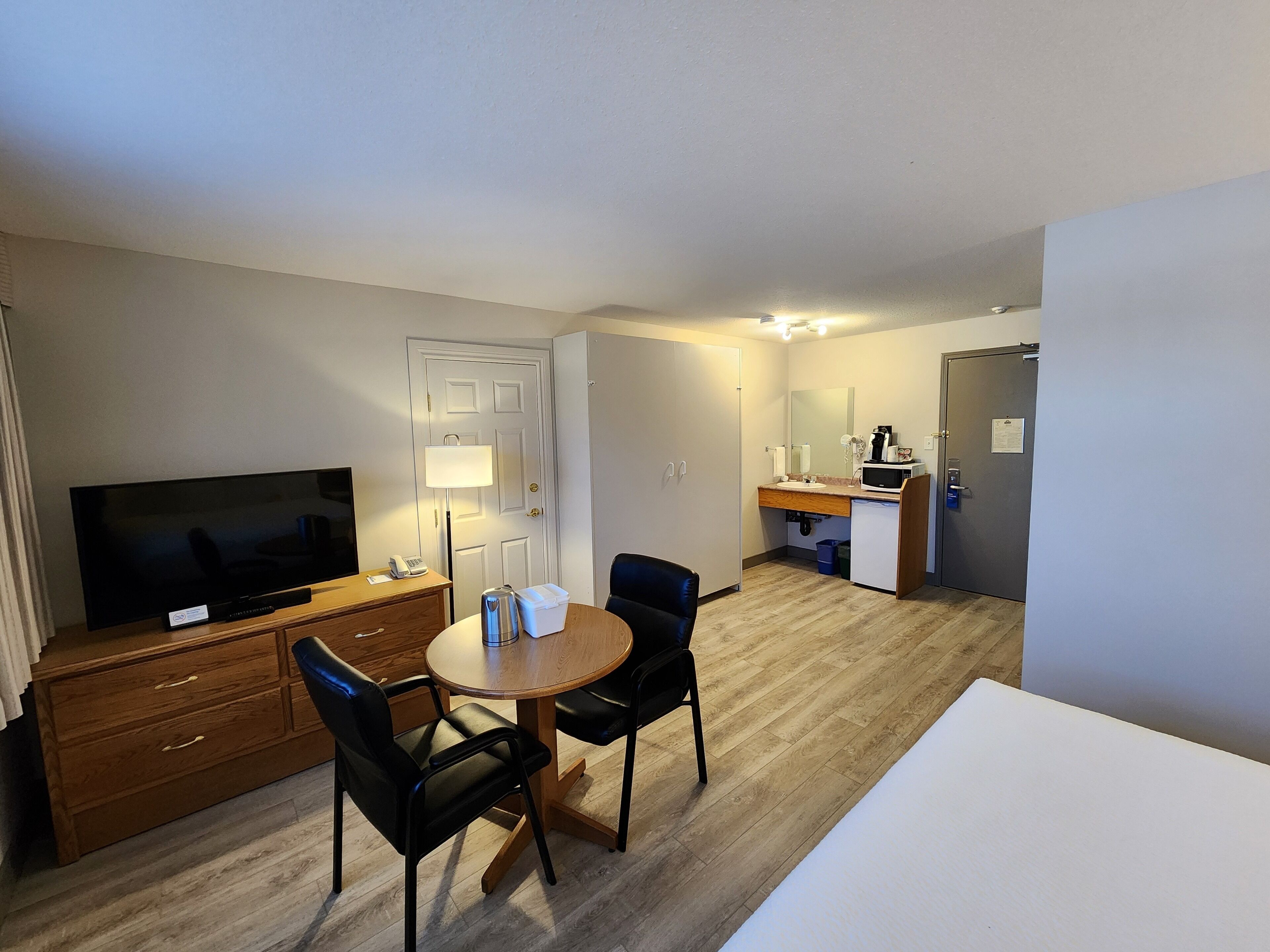 A hotel room with a TV, wooden dresser, two chairs, a round table, a small kitchenette, and a door leading to another room.