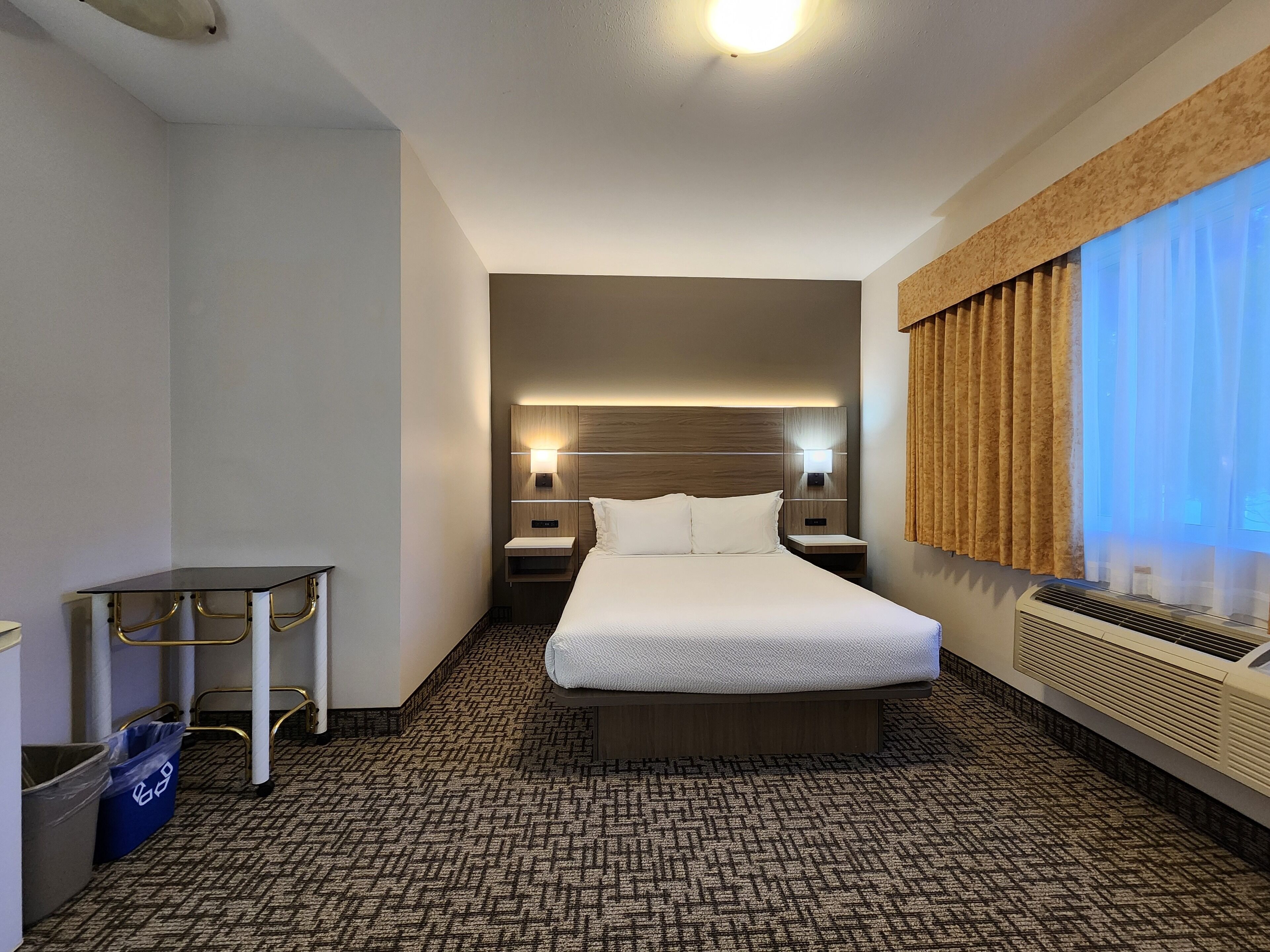 A hotel room with a bed, bedside tables, a desk, a trash bin, and a window with curtains.