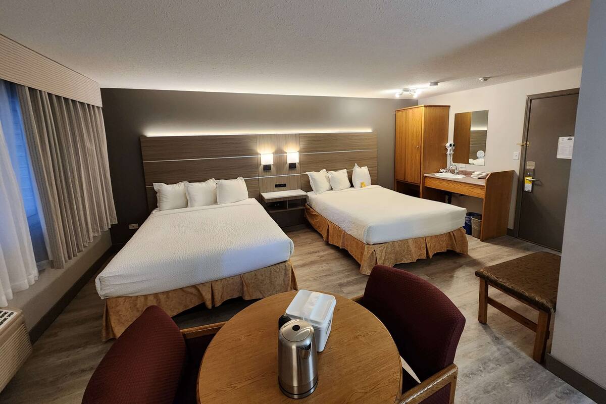 Habitación de hotel con dos camas, una mesa de madera, una silla, una pequeña kitchenette y un baño con espejo y armario.