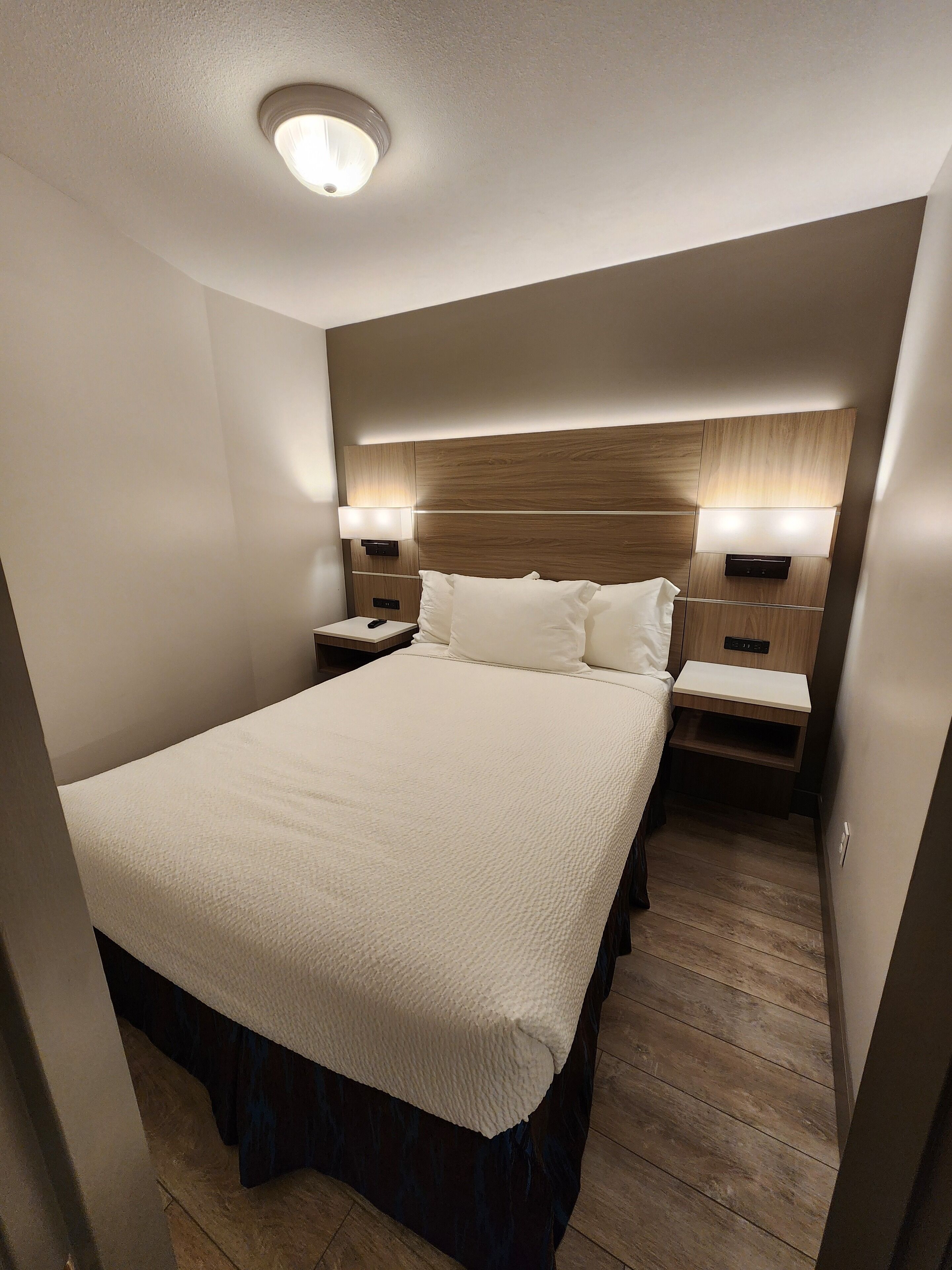 A modern hotel room with a large bed, wooden headboard, bedside lamps, and a built-in nightstand.
