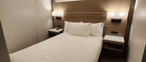 A modern hotel room with a large bed, wooden headboard, bedside lamps, and a built-in nightstand.