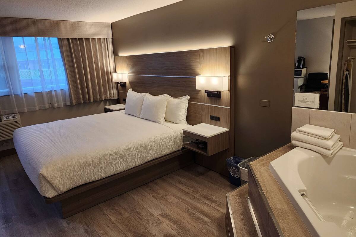 Una habitación de hotel moderna con una cama grande, mesitas de noche, televisión integrada, un baño con bañera y un ventanal con cortinas.