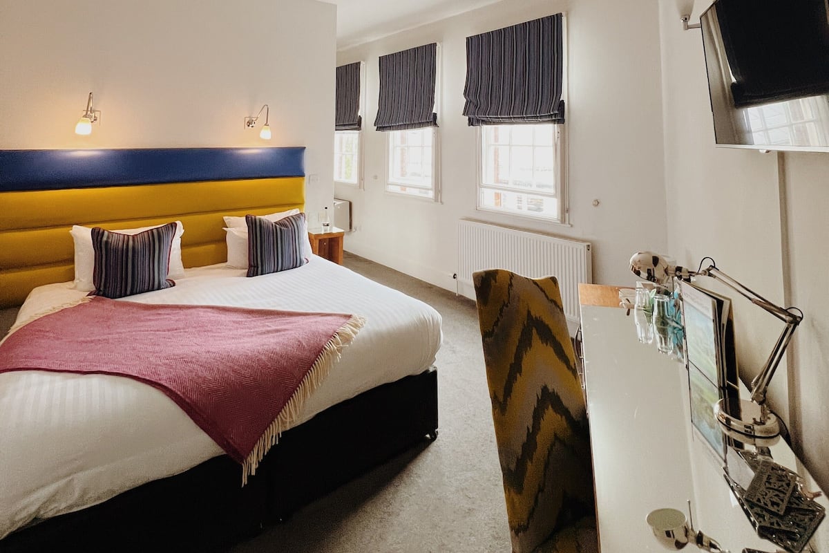A hotel room with a bed, a desk, a chair, a television, a ceiling fan, and windows with curtains.