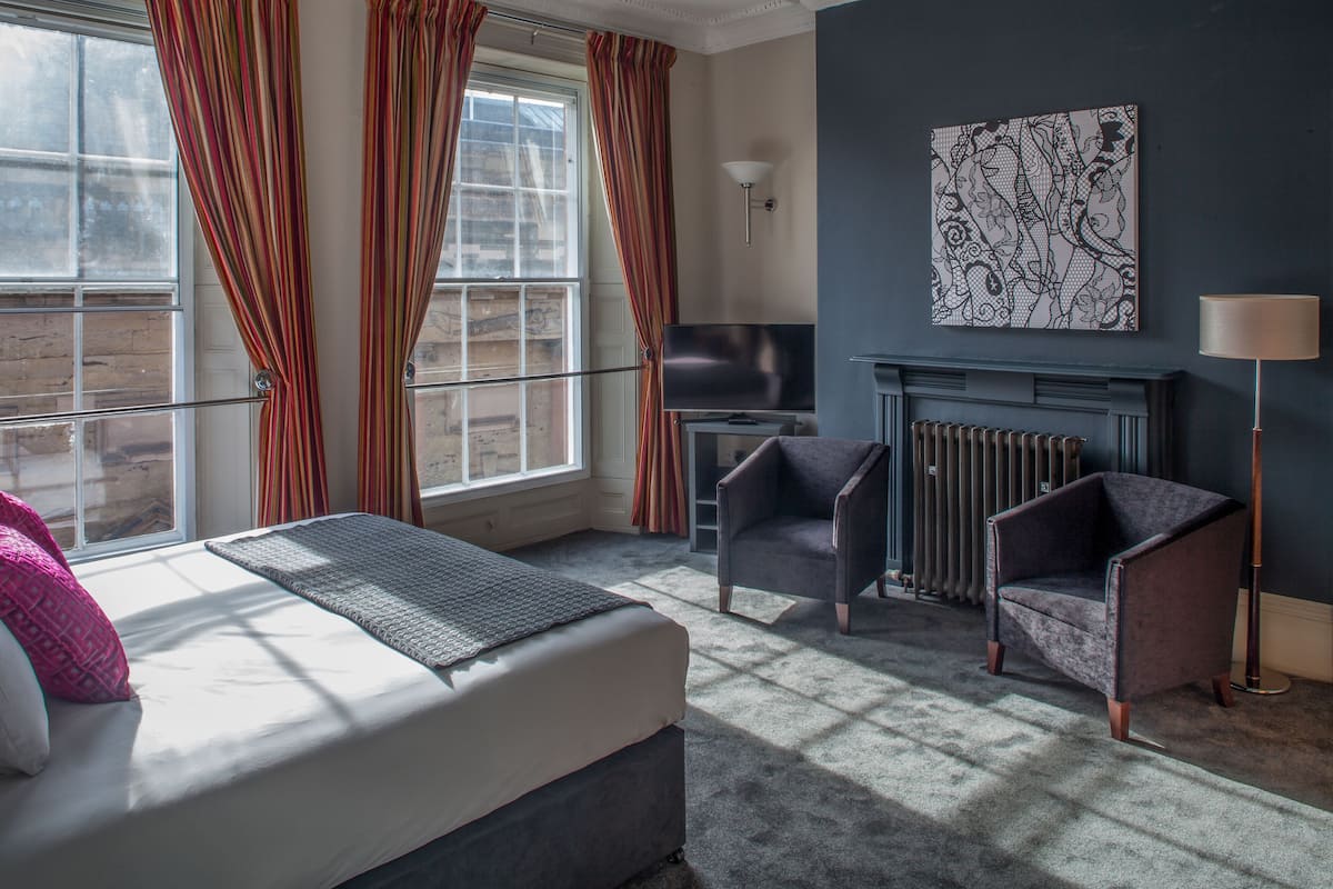 A bedroom with a bed, two armchairs, a TV, a fireplace, and a large window with curtains.
