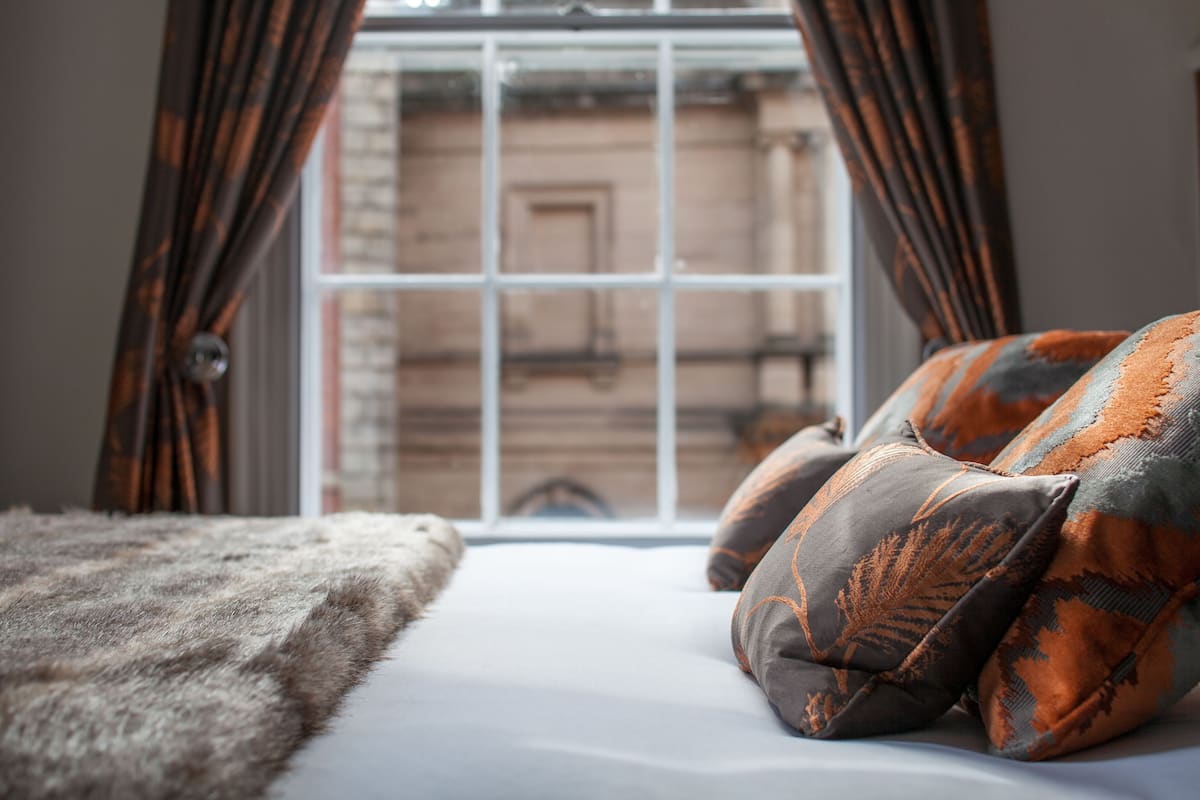 A bed with a fluffy blanket and patterned pillows, a window with curtains, and a view of a building outside.