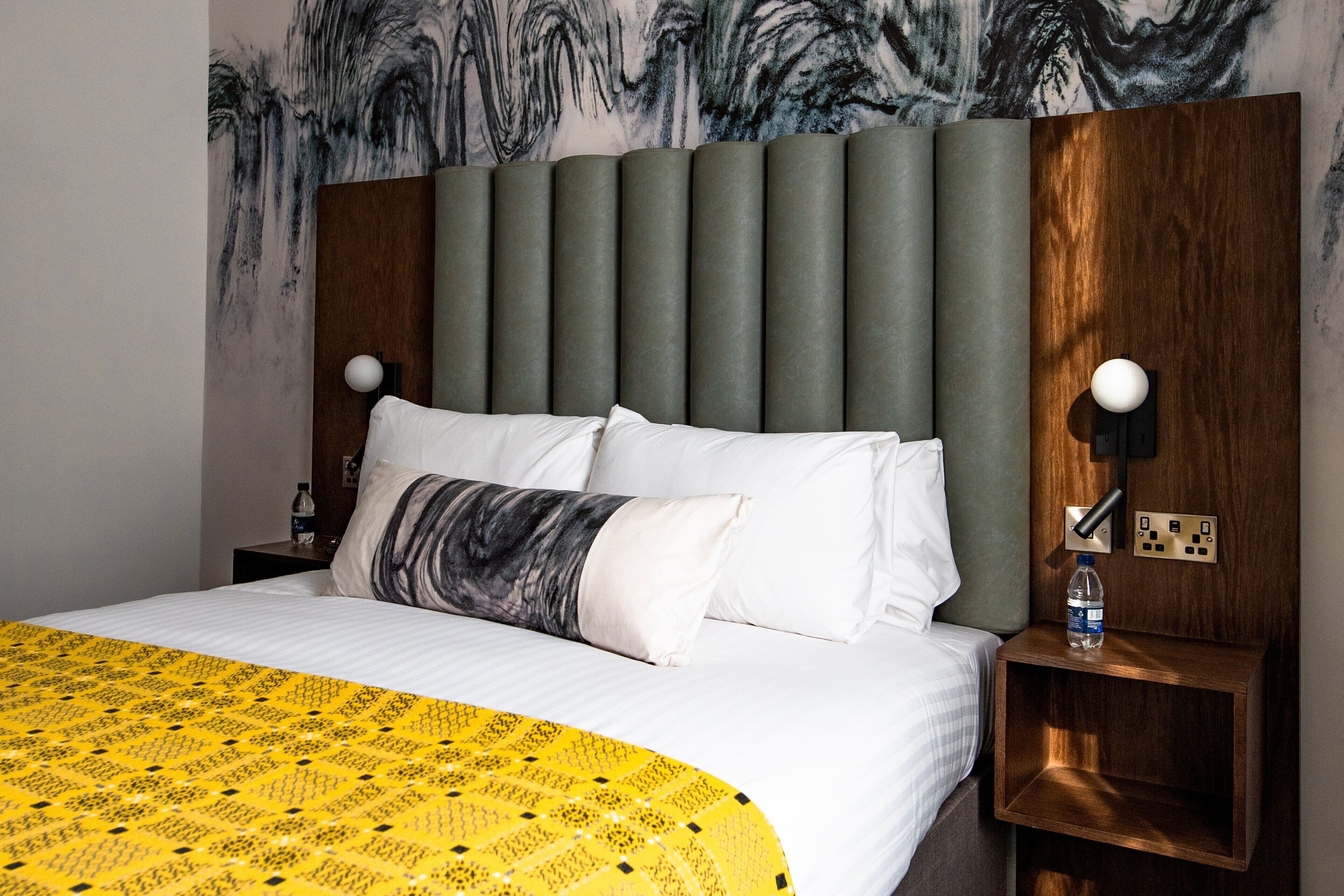A hotel room with a bed, a nightstand, a wall-mounted lamp, and a water bottle on the nightstand.