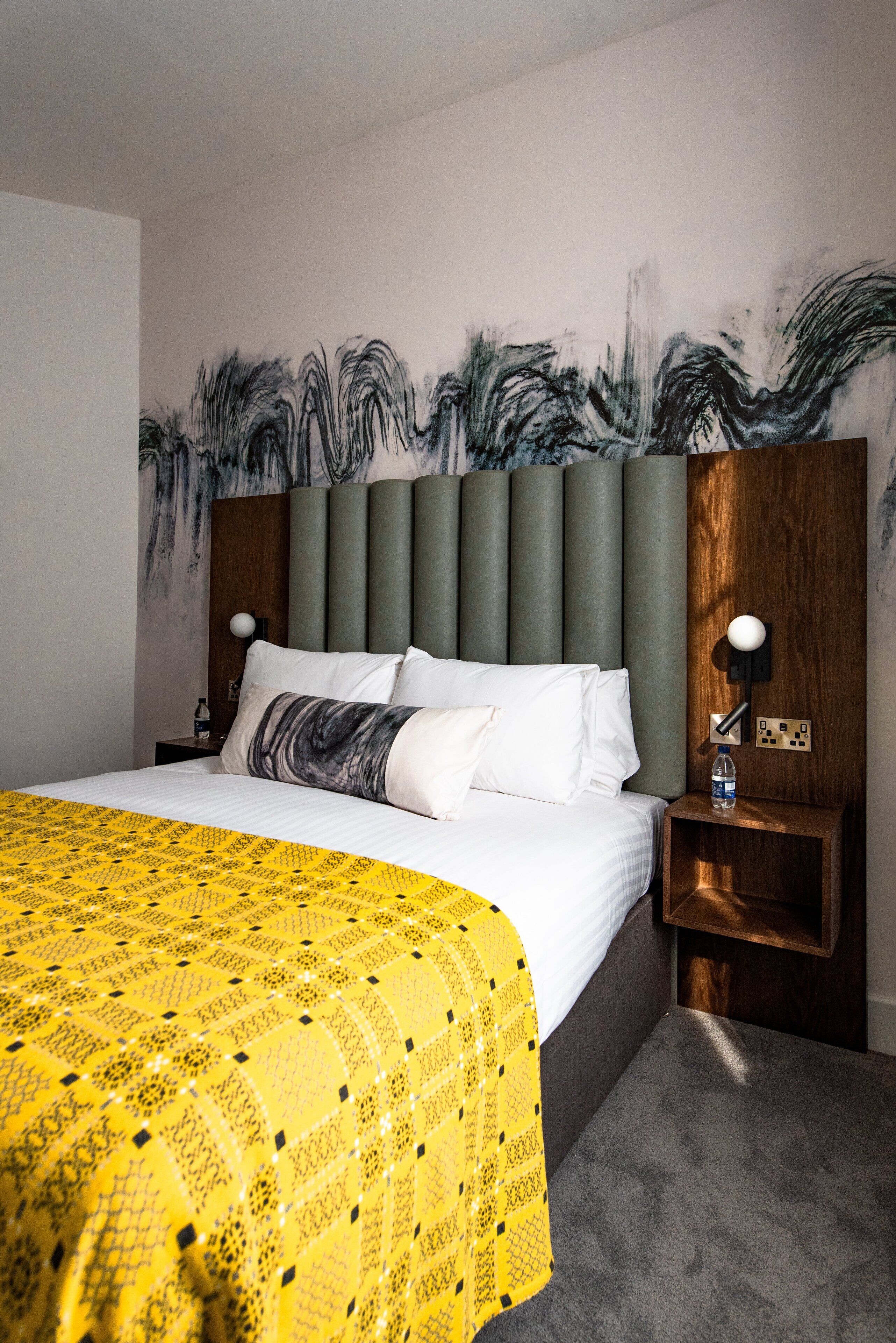 Una habitación de hotel con una cama grande, un cabecero con paneles verticales, una mesita de noche con lámpara y una pared decorada con arte abstracto.