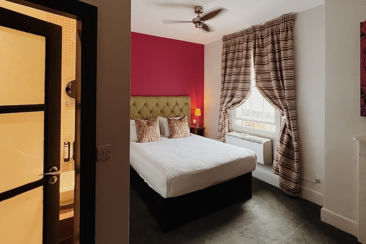 A hotel room with a bed, a ceiling fan, a window with curtains, and a view of the outside.