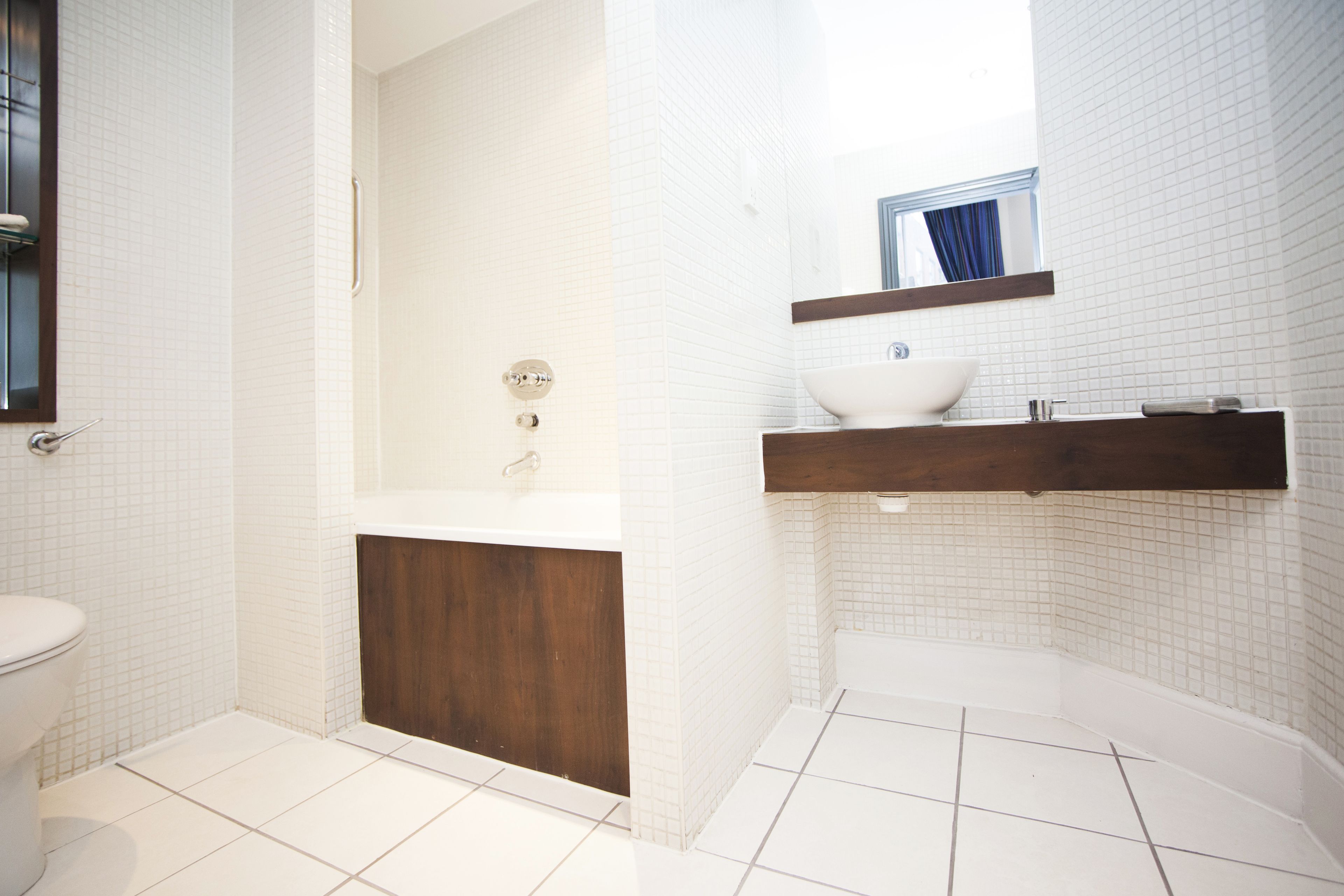 Une salle de bain avec des murs recouverts de carreaux blancs, un meuble-lavabo en bois, un lavabo blanc et une baignoire.