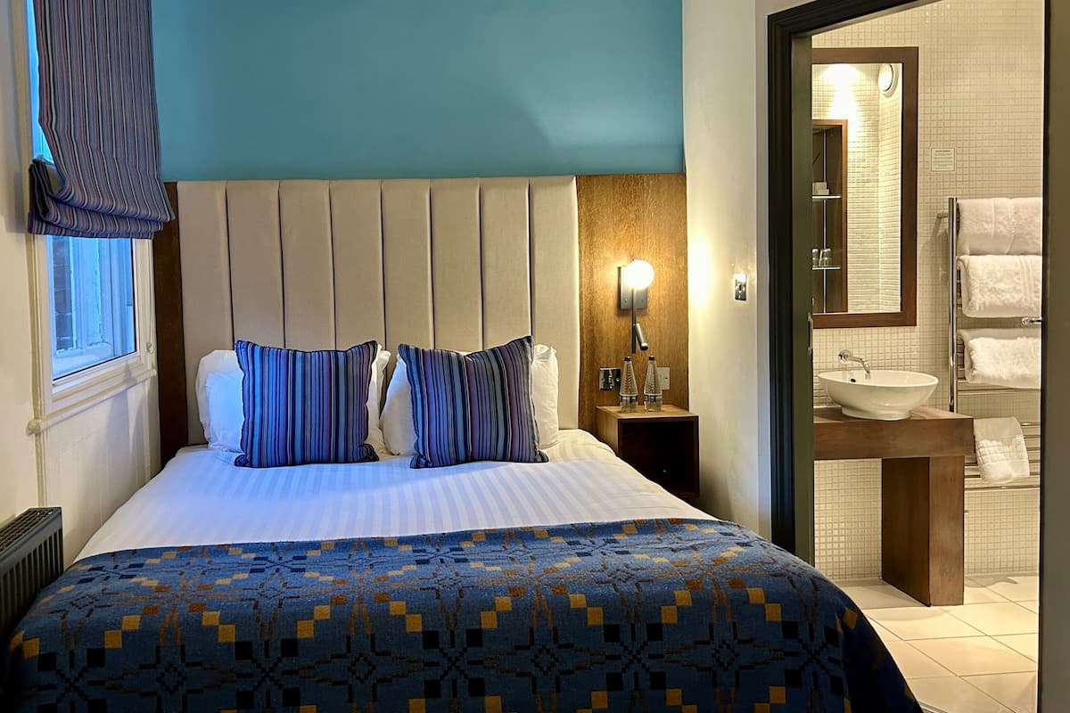 A hotel room with a large bed, a bedside table with a lamp, a bathroom with a sink and towel rack.