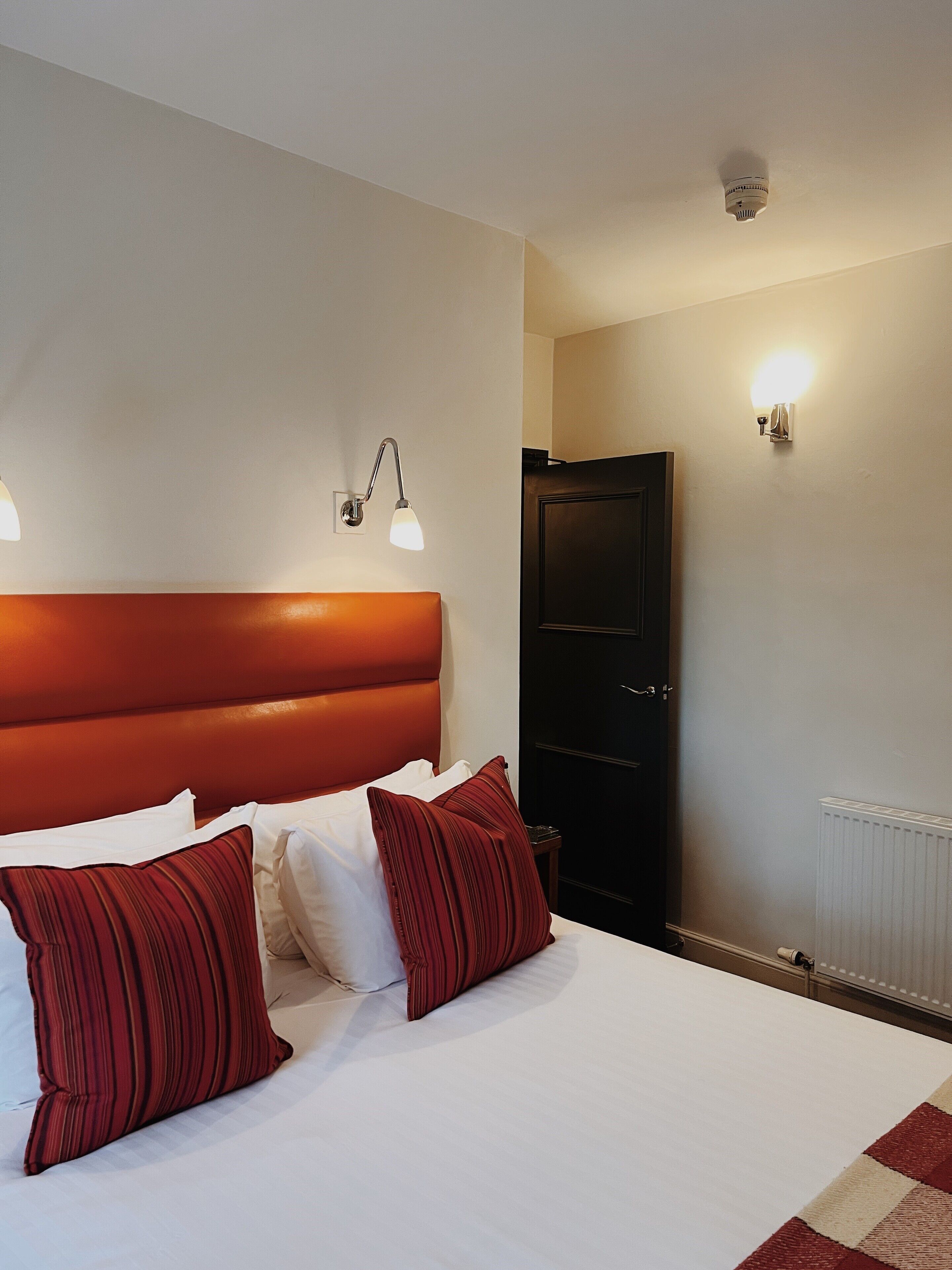 A hotel room with a large bed, two red pillows, a white headboard, and two wall-mounted lights.