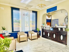 A reception area with a counter, two armchairs, a chandelier, and a large window with curtains.