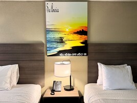 Habitación de hotel con dos camas, una mesita de noche con teléfono y lámpara, y un cartel en la pared con una puesta de sol sobre el Golfo de México.
